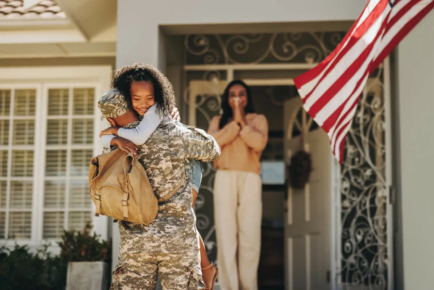 A soldier home from duty hugs his daughter at the front door while his wife watches